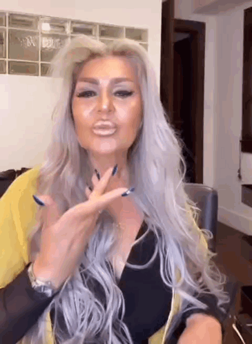 A Woman With Long Gray Hair And Blue Nails Is Sitting At A Table Making A Funny Face . GIF