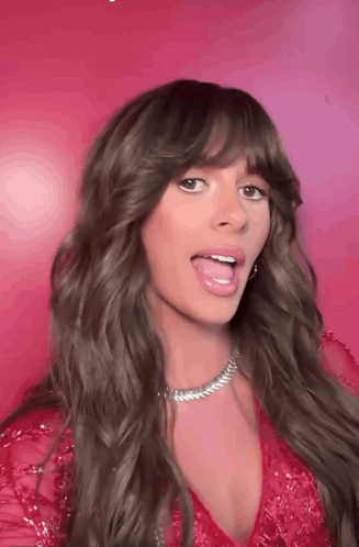 A Woman With Long Hair And Bangs Is Sticking Her Tongue Out In Front Of A Pink Background . GIF