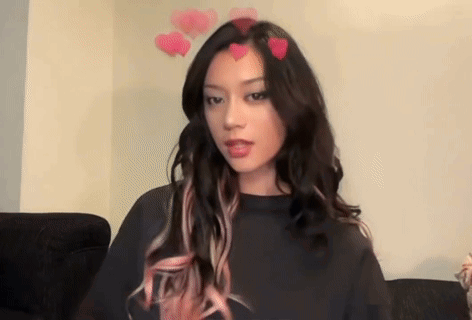 A Woman With Long Hair And Pink Highlights Is Sitting On A Couch With Hearts On Her Head . GIF