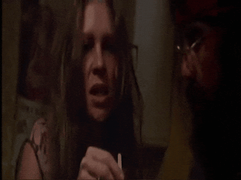 A Woman With Long Hair Is Holding A Cigarette And Talking To A Man With Glasses GIF