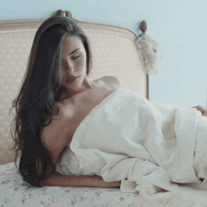 A Woman With Long Hair Is Laying On A Bed With A White Blanket GIF