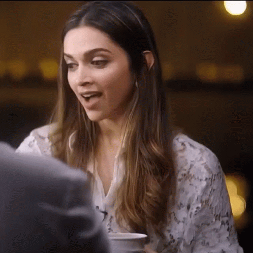 A Woman With Long Hair Is Sitting At A Table With A Cup Of Coffee In Her Hand . GIF