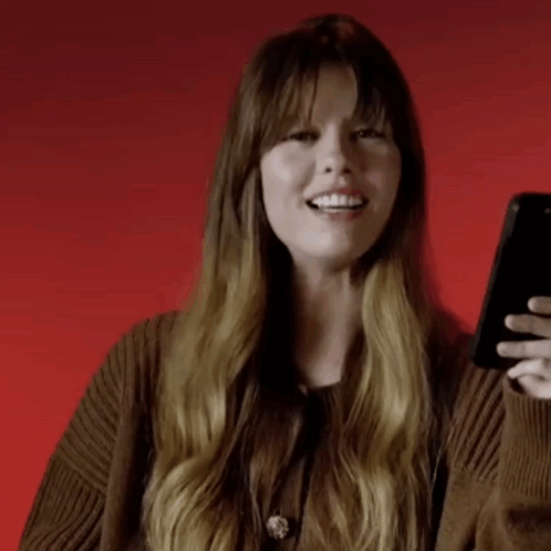 A Woman With Long Hair Is Smiling While Holding A Cell Phone . GIF