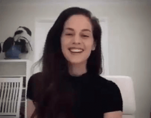 A Woman With Long Hair Is Smiling While Sitting In A Chair In A Room . GIF