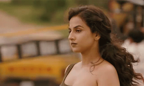 A Woman With Long Hair Is Standing In Front Of A Yellow School Bus . GIF