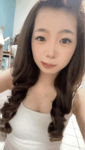 A Woman With Long Hair Is Taking A Selfie In A White Tank Top GIF