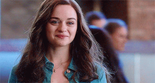 A Woman With Long Hair Is Wearing A Denim Jacket And A Necklace And Smiling . GIF