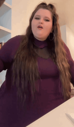 A Woman With Long Hair Is Wearing A Purple Turtleneck And Hoop Earrings GIF