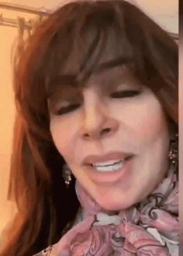 A Woman With Long Hair Is Wearing A Scarf Around Her Neck And Smiling . GIF