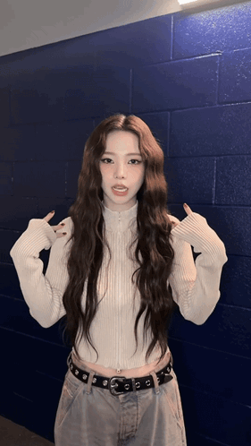 A Woman With Long Hair Wearing A White Sweater And Jeans Stands In Front Of A Blue Brick Wall GIF