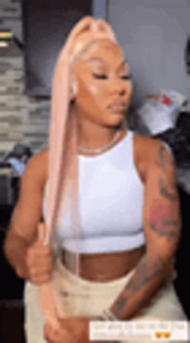 A Woman With Long Pink Hair Is Wearing A White Tank Top And Tan Shorts . GIF