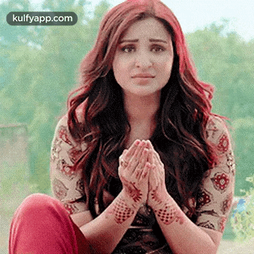 A Woman With Long Red Hair Is Sitting Down With Her Hands Folded In Front Of Her Face . GIF