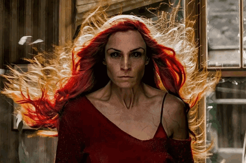 A Woman With Long Red Hair Is Standing In Front Of A Window With Fire Coming Out Of Her Hair GIF