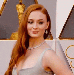 A Woman With Long Red Hair Wearing A Necklace And Earrings Stands In Front Of An Oscar Statue GIF