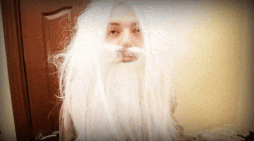 A Woman With Long White Hair And A Beard Looks At The Camera GIF