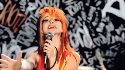 A Woman With Orange Hair Is Singing Into A Microphone In Front Of A Graffiti Wall . GIF