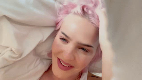 A Woman With Pink Hair Is Laying On A Bed And Smiling GIF