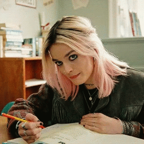 A Woman With Pink Hair Is Sitting At A Desk With A Pencil In Her Hand GIF