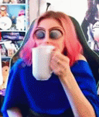 A Woman With Pink Hair Is Sitting In A Chair Drinking From A Mug . GIF