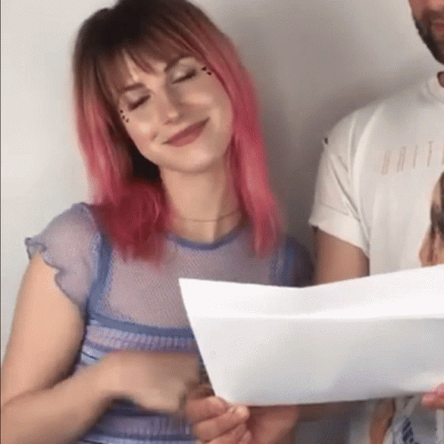 A Woman With Pink Hair Is Smiling While Holding A Piece Of Paper Next To A Man . GIF