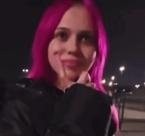 a woman with pink hair is wearing a black jacket and making a face .