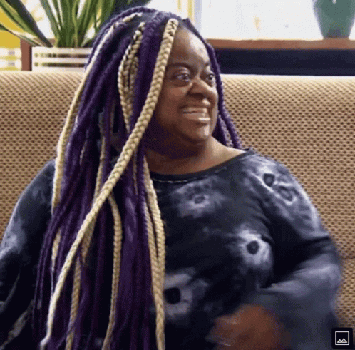 A Woman With Purple Braids Is Smiling And Sitting On A Couch GIF