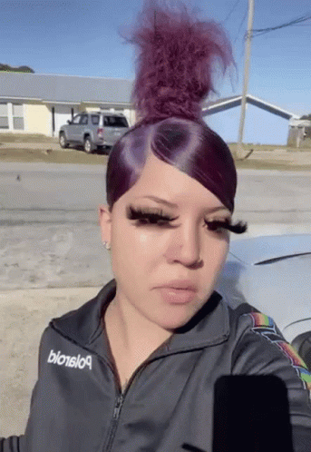 A Woman With Purple Hair And Long Eyelashes Is Standing In Front Of A Car In A Parking Lot . GIF