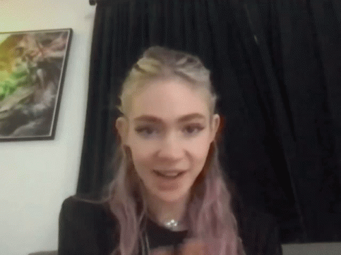 A Woman With Purple Hair Is Sitting In Front Of A Black Curtain And A Picture On The Wall . GIF