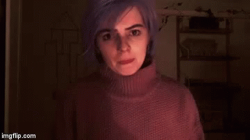 A Woman With Purple Hair Is Wearing A Turtleneck Sweater And Standing In A Dark Room . GIF