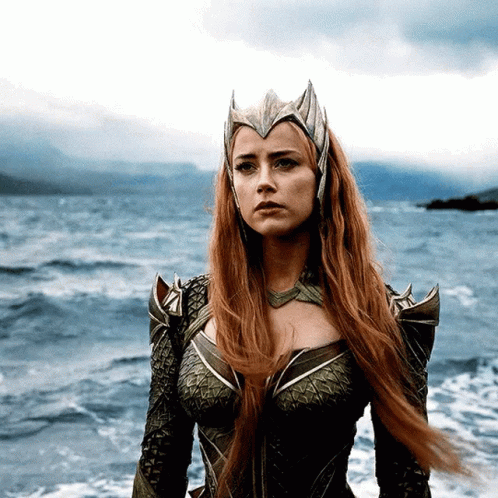 A Woman With Red Hair And A Crown On Her Head Stands In Front Of A Body Of Water GIF