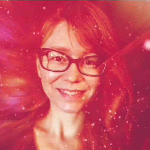 A Woman With Red Hair And Glasses Smiles In A Pink Background GIF