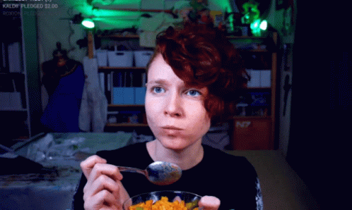 A Woman With Red Hair Is Eating A Bowl Of Food With A Spoon In Front Of A Sign That Says N7 On It GIF