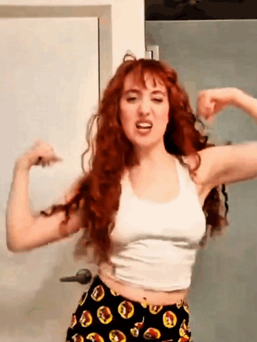 A Woman With Red Hair Is Flexing Her Muscles In Front Of A Door . GIF