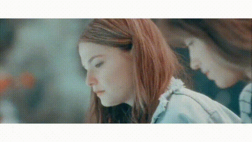 A Woman With Red Hair Is Looking Down At Something While Another Woman Looks At Her Reflection In The Mirror . GIF