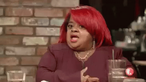 A Woman With Red Hair Is Sitting At A Table In Front Of A Brick Wall And Making A Funny Face . GIF