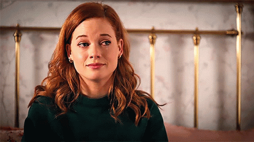 A Woman With Red Hair Is Sitting In Front Of A Bed . GIF