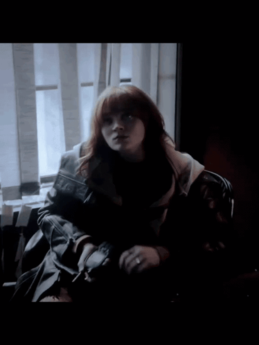 A Woman With Red Hair Is Sitting In Front Of A Window In A Dark Room GIF