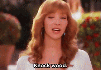 A Woman With Red Hair Is Standing In Front Of A Bush And Says `` Knock Wood '' . GIF