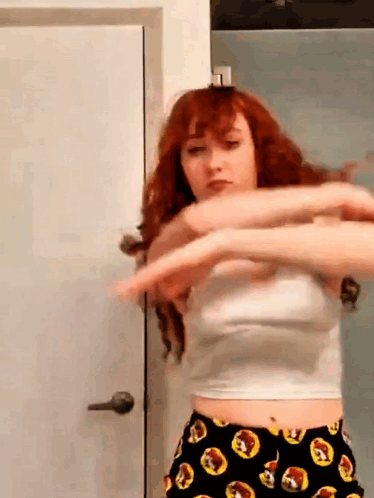 A Woman With Red Hair Is Standing In Front Of A Door Wearing A White Crop Top And Black Shorts . GIF