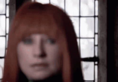 A Woman With Red Hair Is Standing In Front Of A Window And Looking At The Camera . GIF
