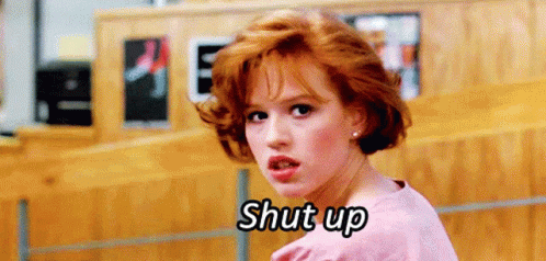 A Woman With Red Hair Is Standing In Front Of A Wooden Wall And Saying Shut Up . GIF