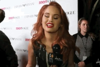 A Woman With Red Hair Is Talking Into A Microphone While Standing In Front Of A Wall That Says Teenvogue . GIF