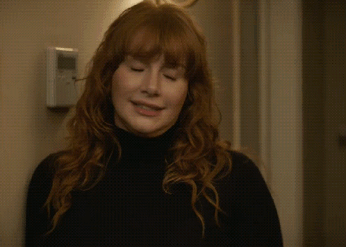 A Woman With Red Hair Is Waving Her Hand In Front Of A Door GIF