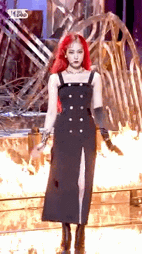A Woman With Red Hair Is Wearing A Black Dress And Standing In Front Of A Fire . GIF