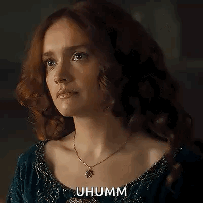 A Woman With Red Hair Is Wearing A Necklace And A Blue Dress And Looking At The Camera . GIF