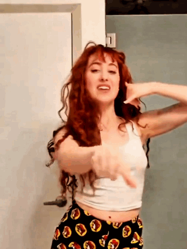A Woman With Red Hair Is Wearing A White Tank Top And Black Pants GIF