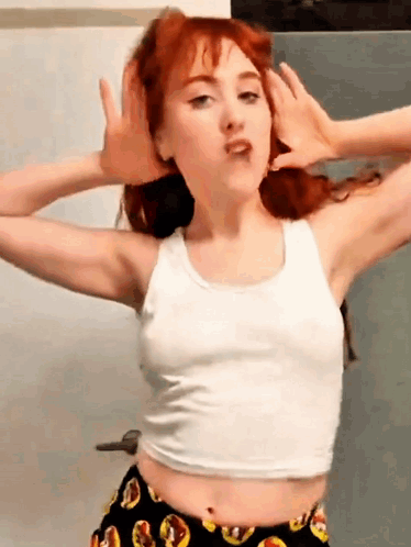 A Woman With Red Hair Is Wearing A White Tank Top And Black Shorts GIF