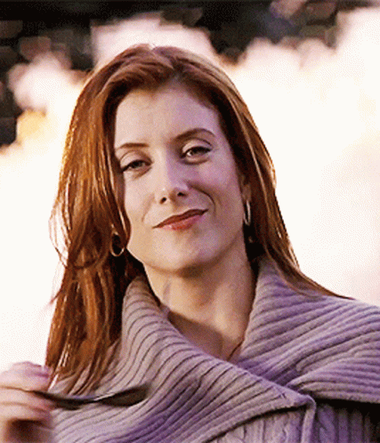 A Woman With Red Hair Wearing A Grey Sweater And Hoop Earrings GIF