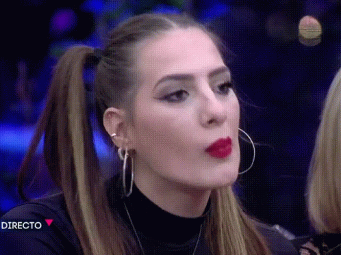 A Woman With Red Lipstick And Hoop Earrings Is Making A Face In Front Of A Directo Sign GIF