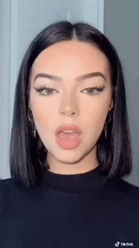 A Woman With Short Black Hair And Earrings Is Wearing A Black Sweater And Making A Funny Face . GIF